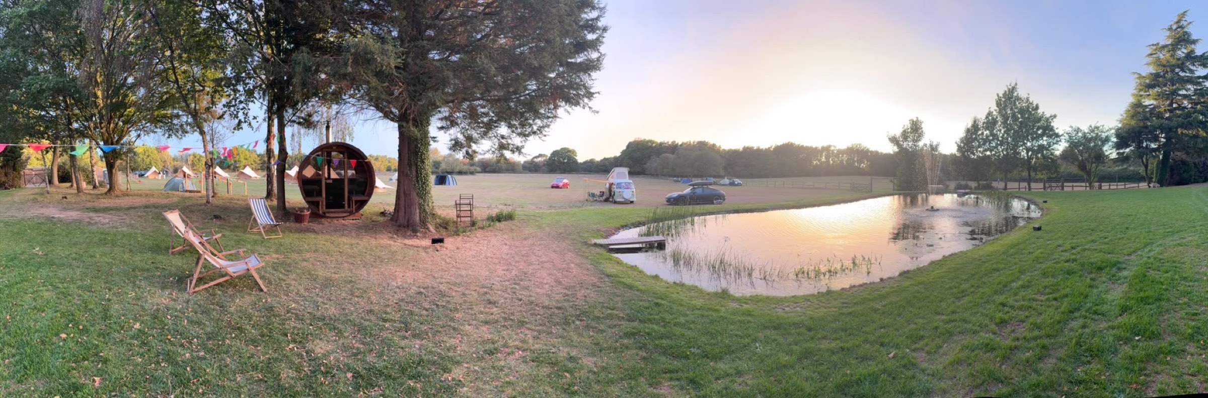 Panorama of the lawn at sunset: the barrel sauna, deckchairs, the tents and the pond