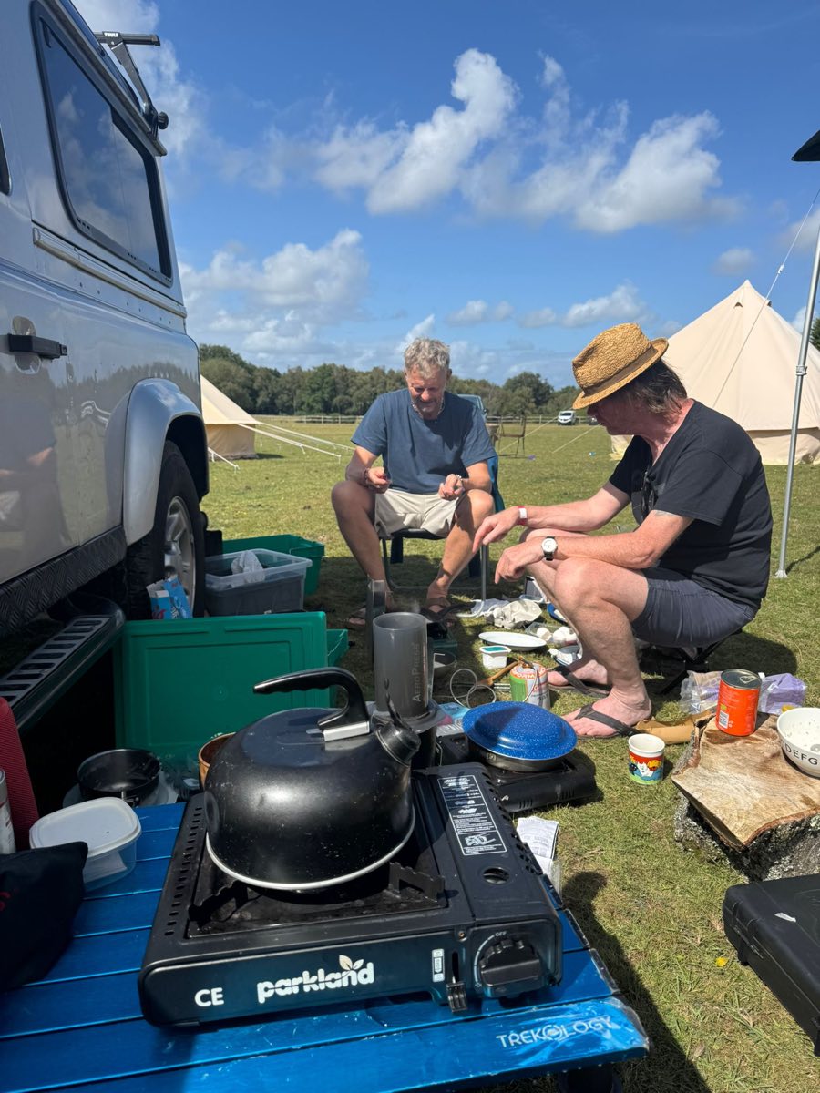 Two campers cooking on a stove beside a Land Rover, with bell tents in the field behind