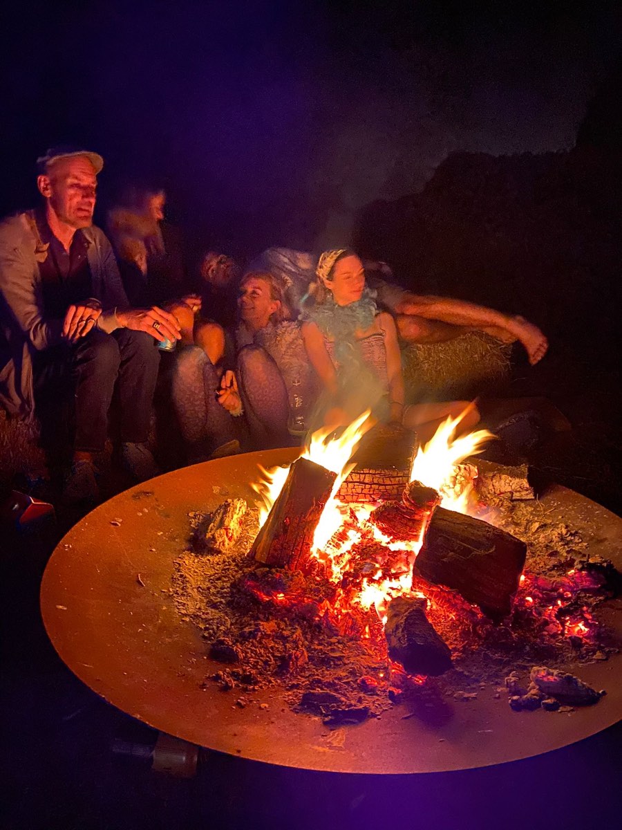Friends on hay bales round a big fire bowl at night, logs blazing