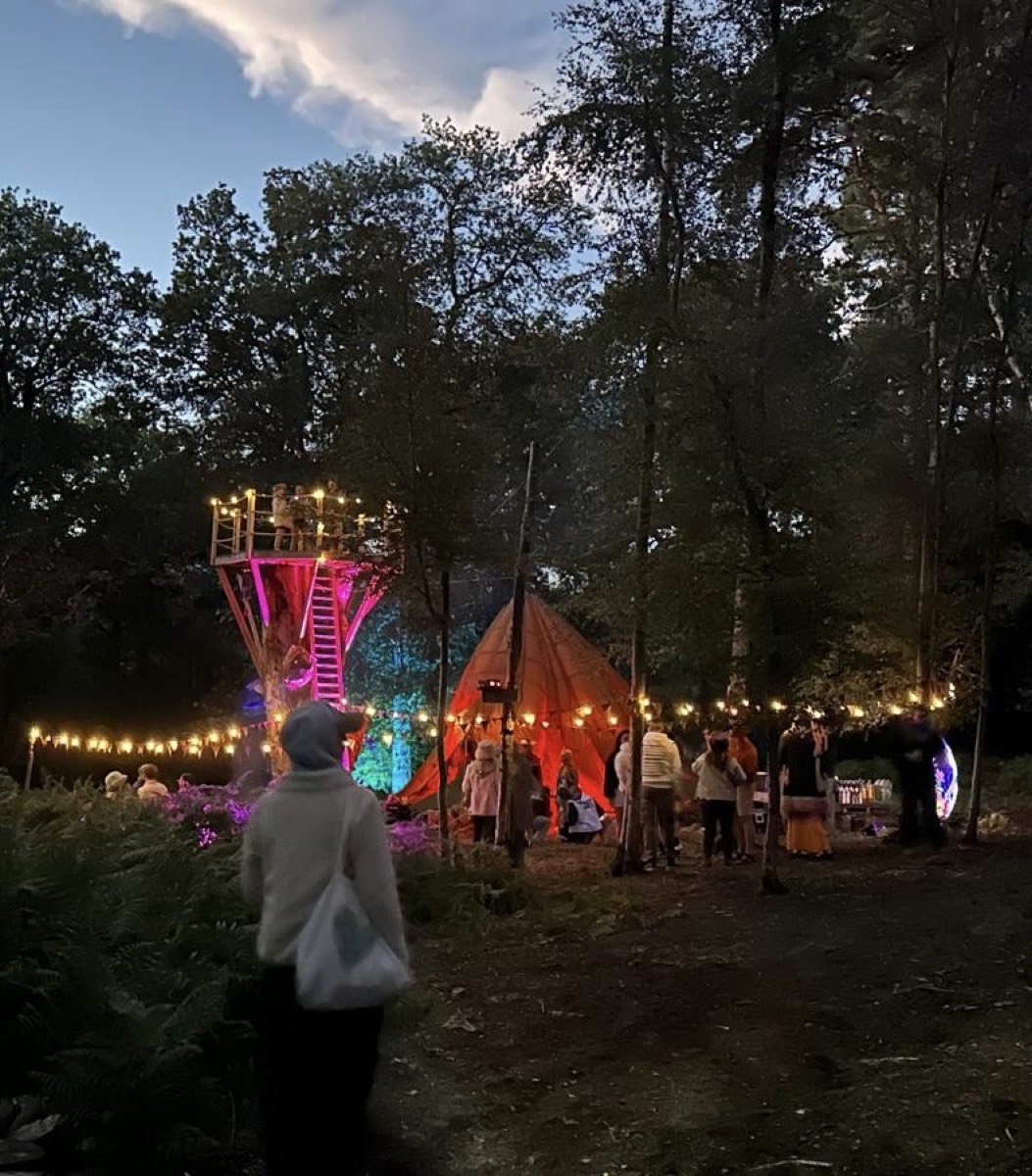 Dusk in the woods: a lit tower and tipi with fairy lights and people gathering underneath