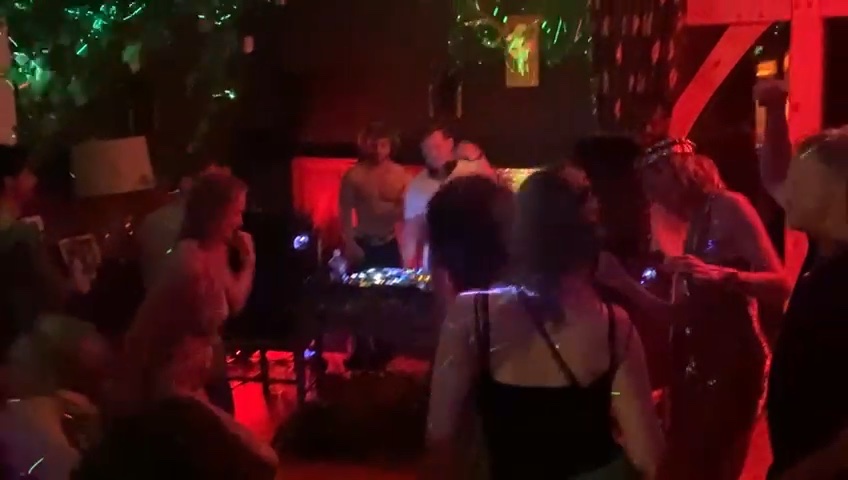 A packed dance floor in red light with two DJs behind the decks
