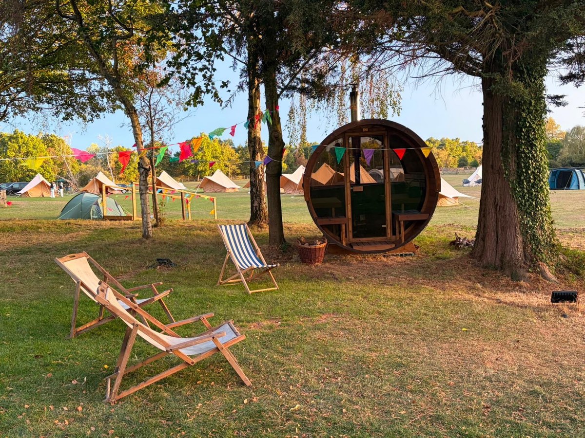 The barrel sauna under the trees, with deckchairs on the lawn and bell tents beyond